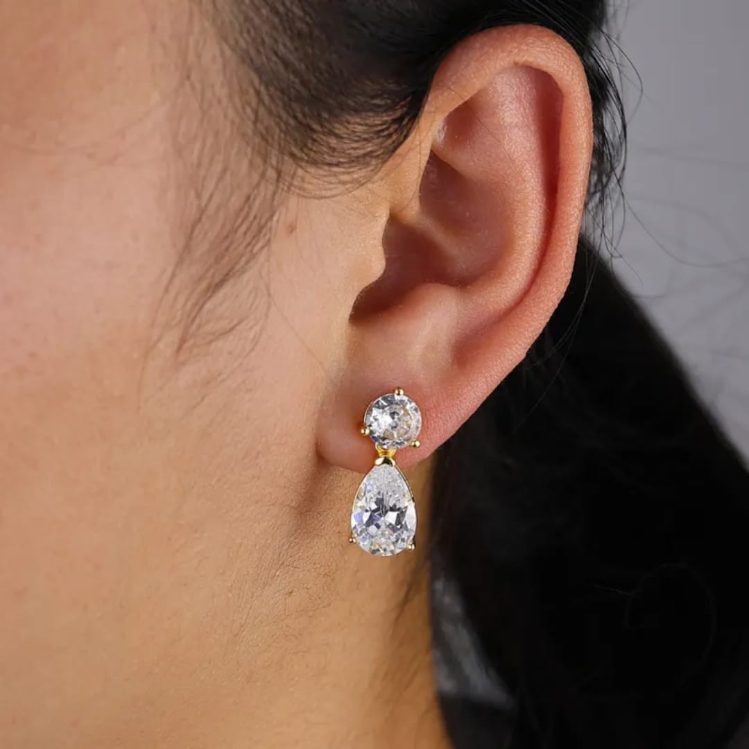 TITLIYA gold-plated drop earrings featuring a round and pear-cut diamond-like stone, worn on a woman's ear.