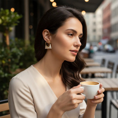 Woman wearing TITLIYA cute bag-shaped earrings with a gold hoop and cream-colored base at an outdoor cafe.