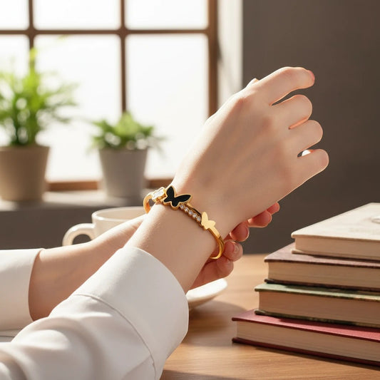 TITLIYA gold-tone bracelet with black butterfly and floral accents, worn on a wrist near books and a window.