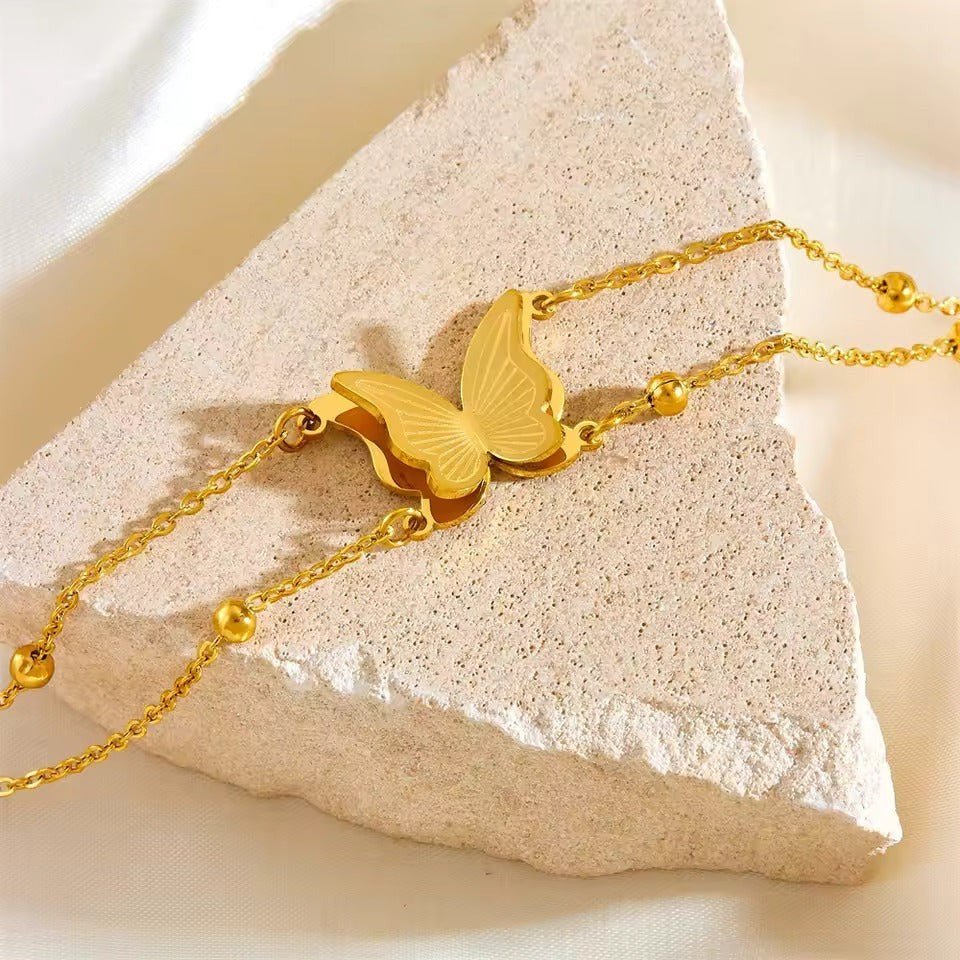 TITLIYA gold-tone butterfly charm bracelet with delicate chain and polished bead accents on a textured stone surface.