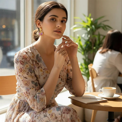 Woman wearing TITLIYA gold-plated butterfly earrings with purple stones, styled with a floral dress in a cafe.