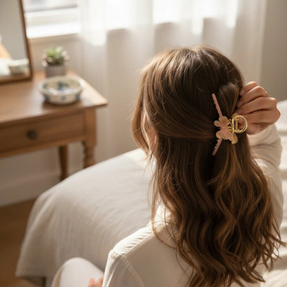 TITLIYA butterfly hair claw in pink and cream enamel with gold finish worn by a woman in a bright bedroom setting.