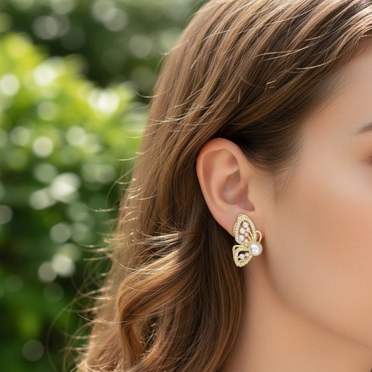 TITLIYA gold butterfly wing stud earrings with pearl accents worn by a woman against a soft green outdoor background