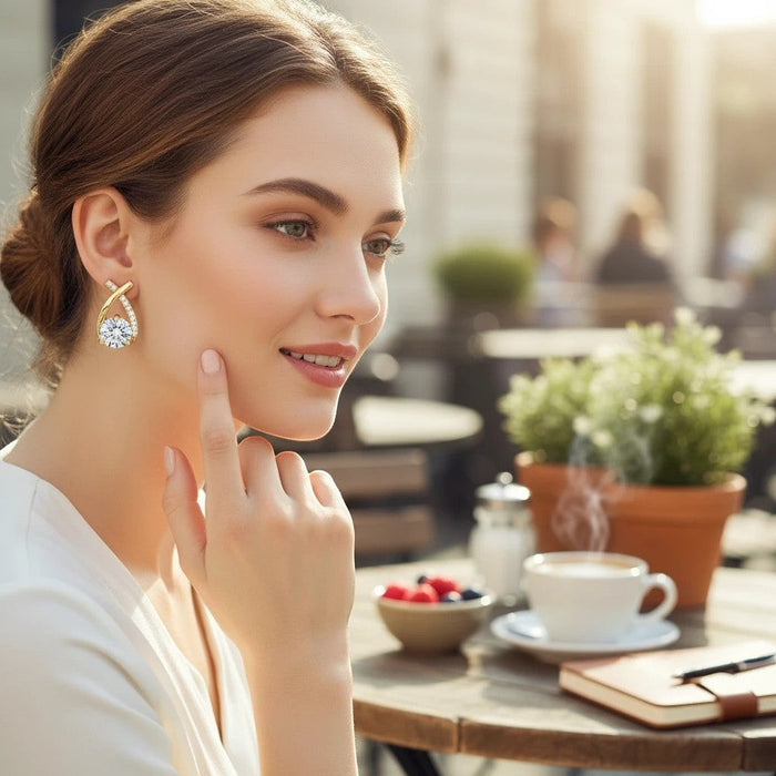 Woman wearing a gold crystal cross-shaped earring by TITLIYA, styled for an elegant daily look at an outdoor cafe.