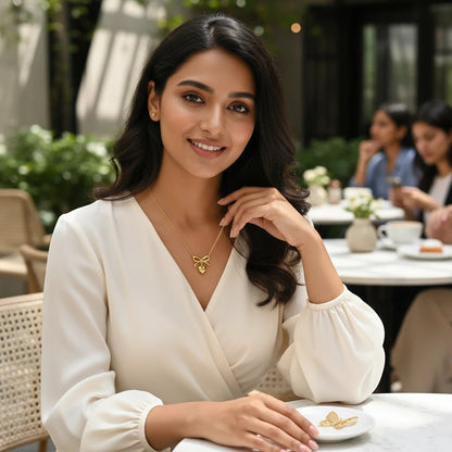 Woman wearing a TITLIYA gold heart and bow pendant necklace with a white blouse in a cafe setting.