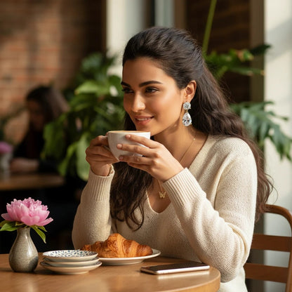 Woman wearing TITLIYA gold-plated drop earrings with pear-shaped diamonds while drinking coffee in a cafe.