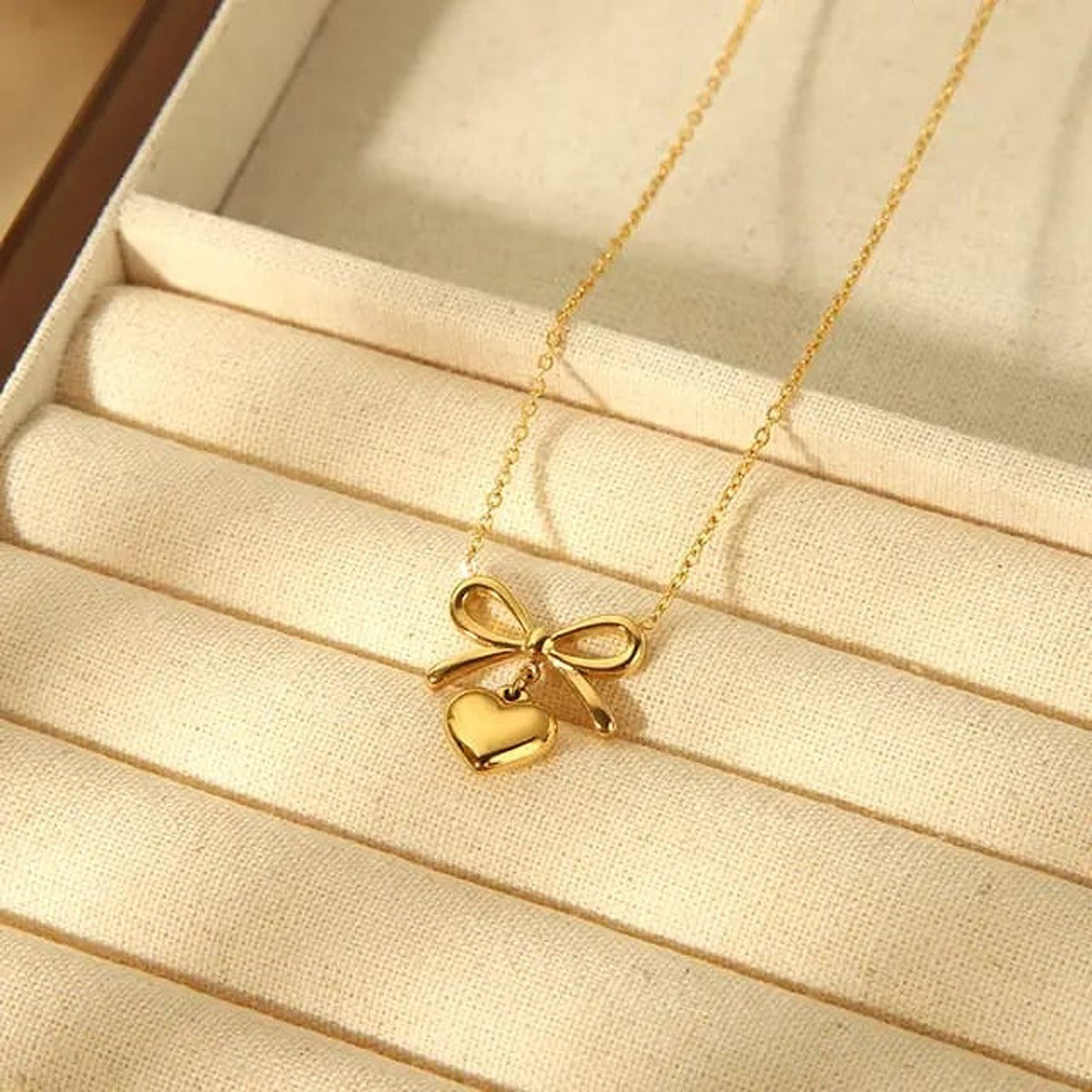 TITLIYA golden necklace featuring a delicate bow pendant with a dangling heart charm on a fine chain in a jewelry box.