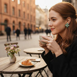 Woman wearing TITLIYA sea green enamel flower stud earrings with gold-tone center at a cafe table.